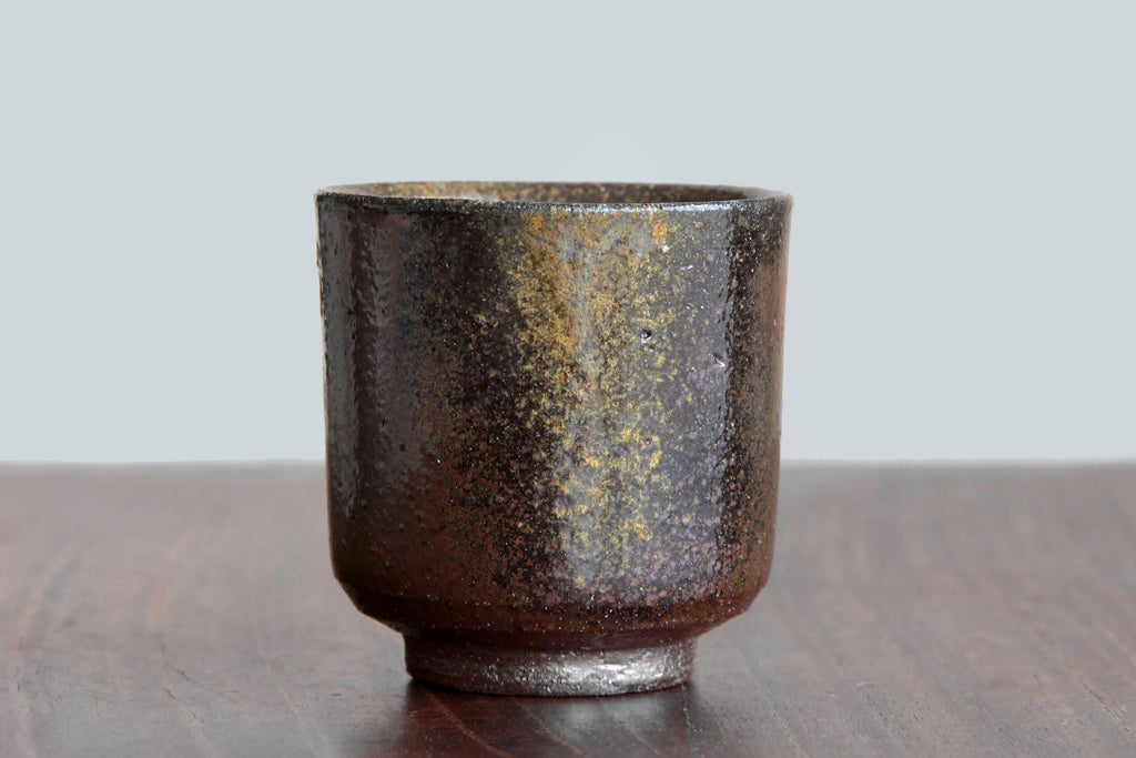 Bizen ware yunomi tea cup with natural ash glaze patterns in dark brown and golden tones, displayed on a wooden surface