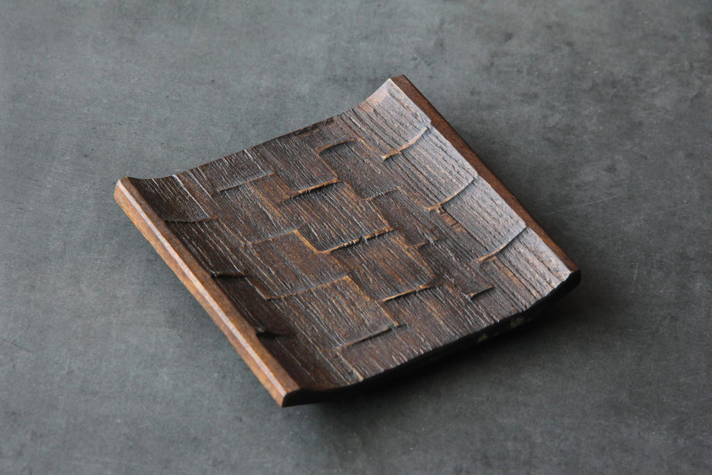 Japanese handcrafted wooden plate with carved geometric texture and dark brown finish, displayed on a gray surface.