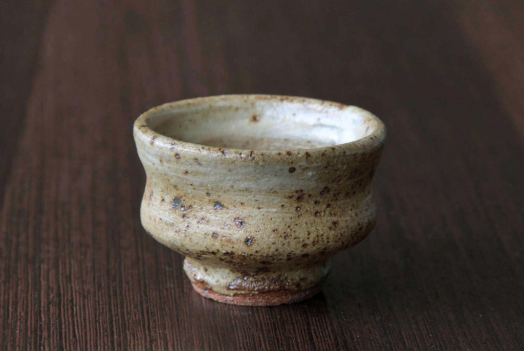Single Japanese pottery sake cup with textured beige glaze
and speckled surface.