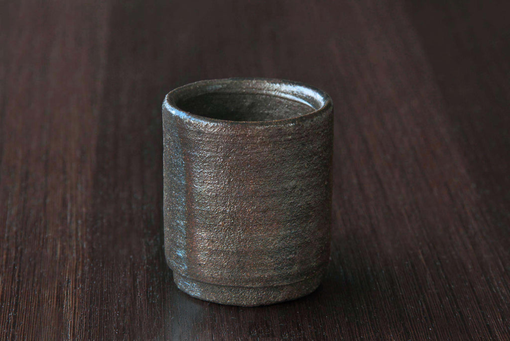 Bizen ware tea cup with cylindrical form and rustic earthy surface, displayed upright on a dark wooden table.
