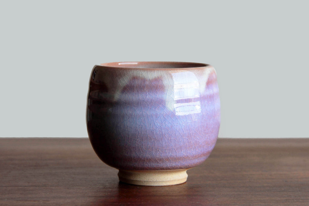 Side view of a Hagi ware yunomi tea cup with vivid purple and pink glaze on a wooden
surface