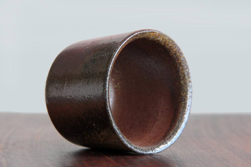Unglazed Japanese pottery yunomi tea cup with dark brown natural clay texture, placed on its side on a wooden surface