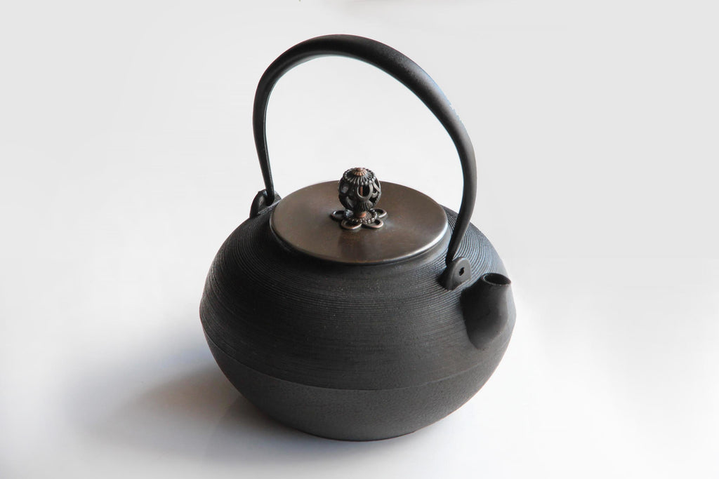 A traditional Japanese Nambu cast iron teapot with a rounded body, elegant curved handle, and a finely crafted copper lid featuring an intricate floral knob, displayed on a clean white surface.