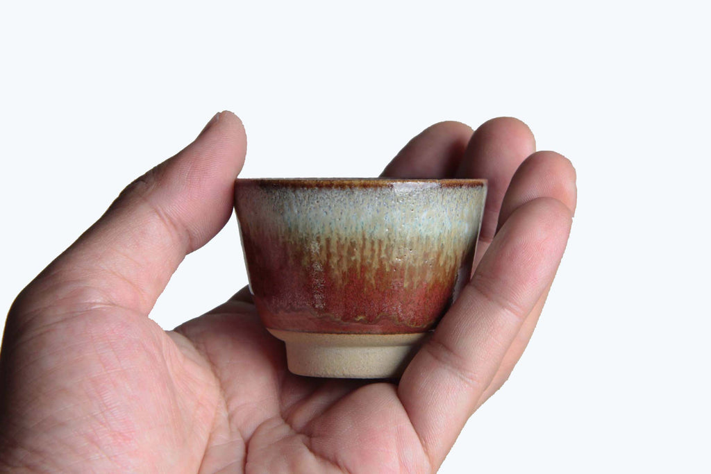 Hand holding a small Japanese sake cup with red and ash glaze.