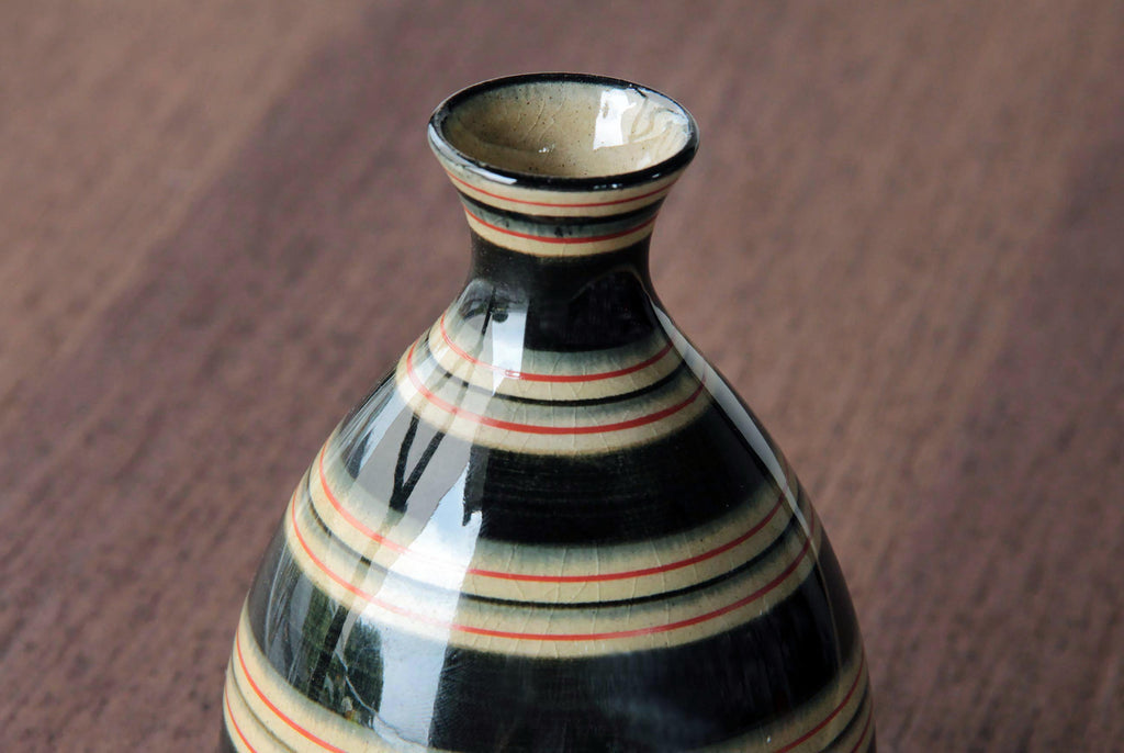 Tokkuri (Japanese sake bottle) with black, ivory, and red horizontal stripes in Hasami-yaki style, placed on wooden table.