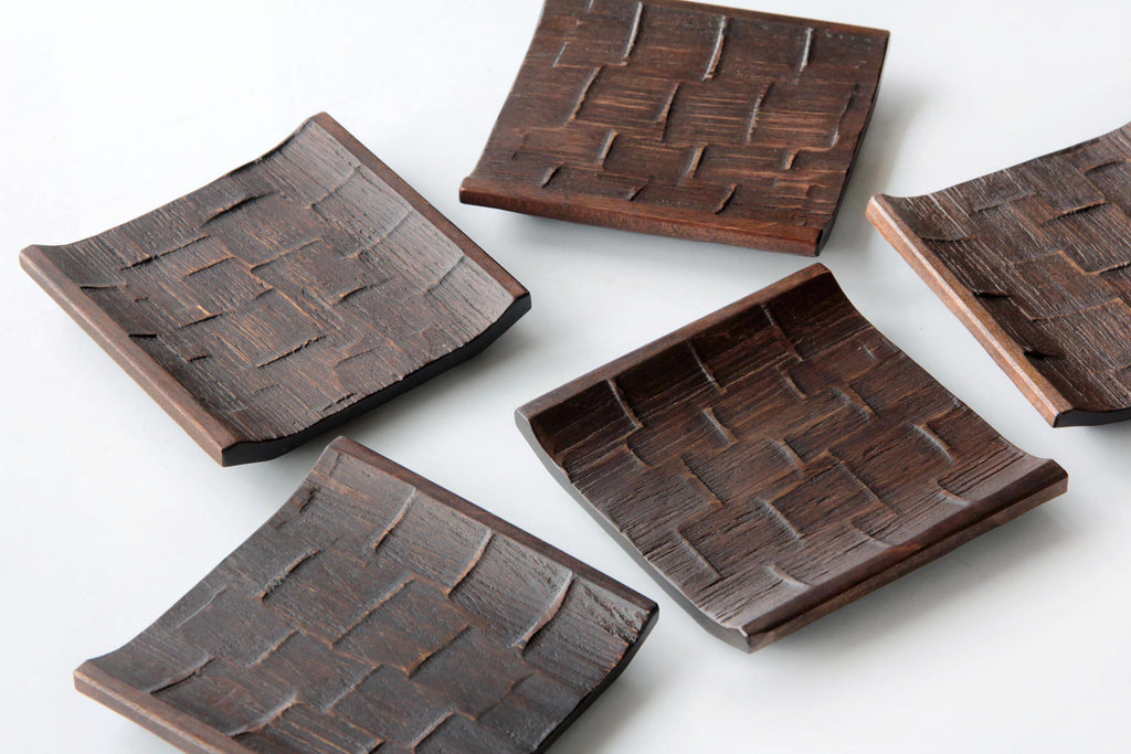 Hand-carved Japanese wooden square plates with a textured lattice pattern, arranged in a scattered layout on a white surface