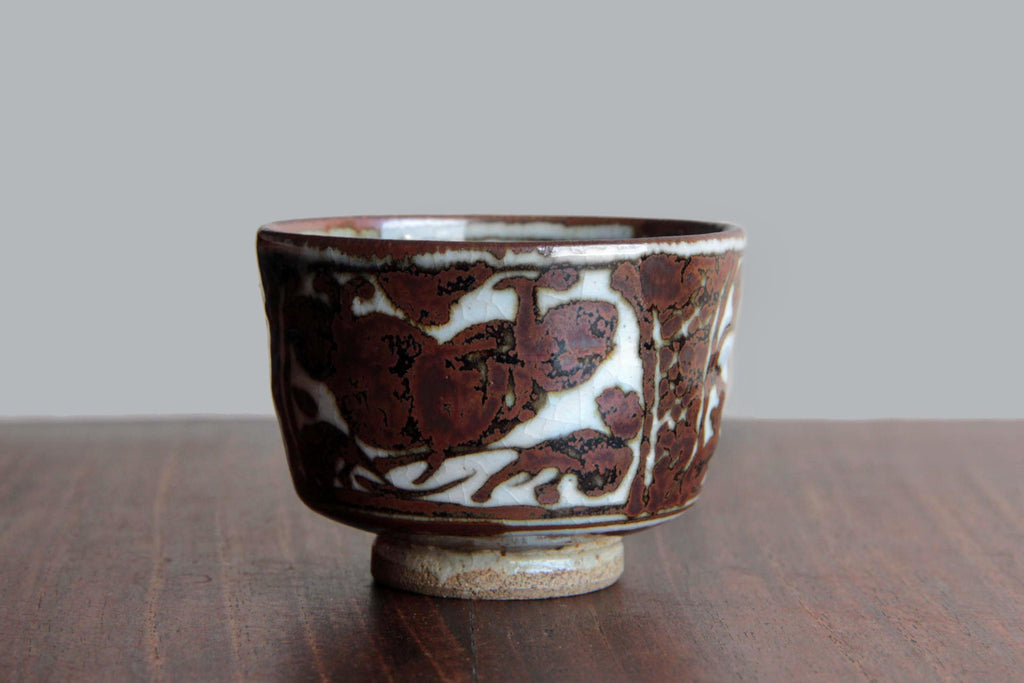 Side view of a Kyoto-style pottery cup with floral and geometric motifs painted in reddish-brown on white base