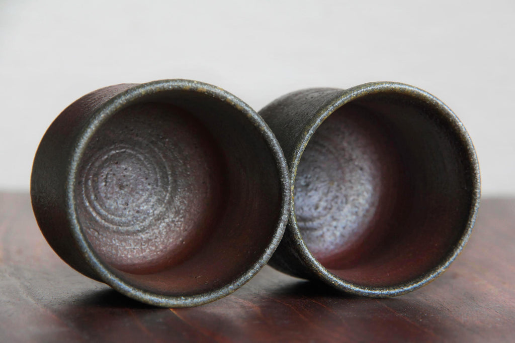 Pair of small Bizen ware tea cups with rough textured surfaces and natural ash glaze effects, placed side by side on a wooden table.