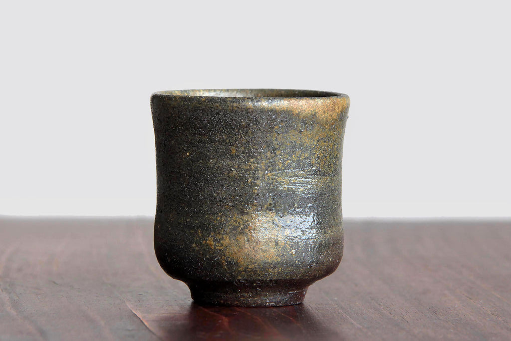 Small Bizen ware tea cup with rough dark brown surface and natural kiln-fired textures, displayed on a wooden table against a light background