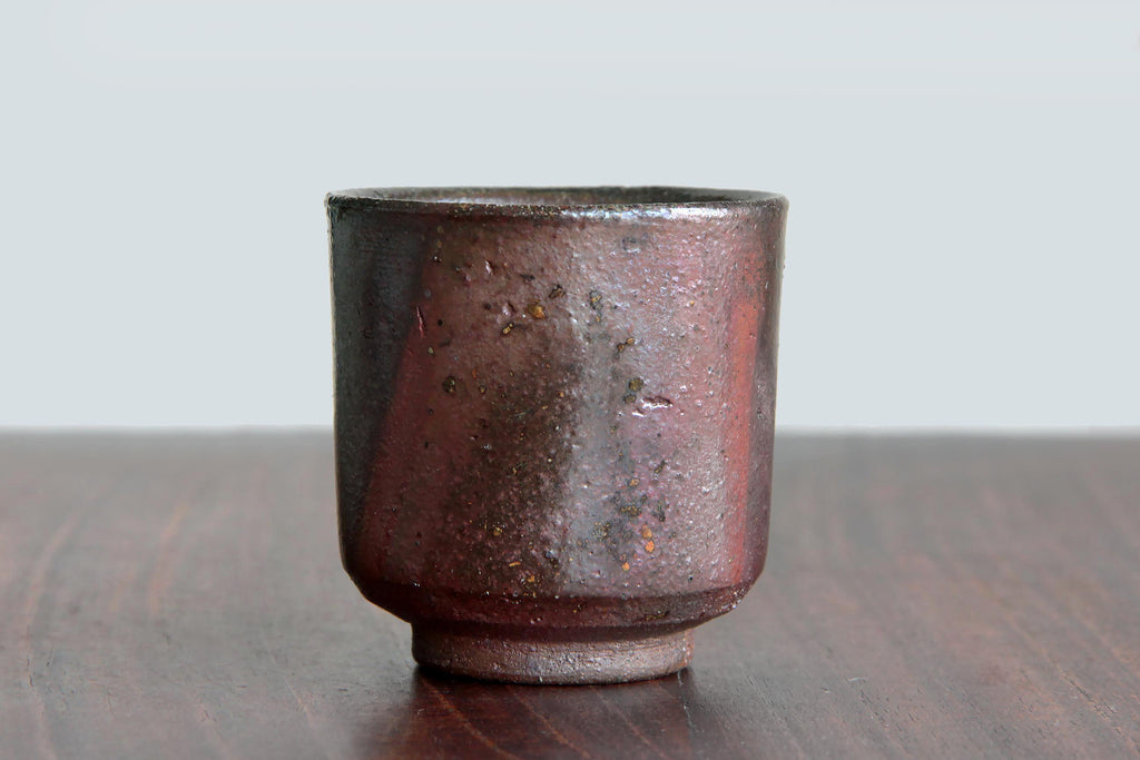 Unglazed Japanese pottery cup with natural ash glaze effects and earthy reddish-brown surface, displayed on a wooden table